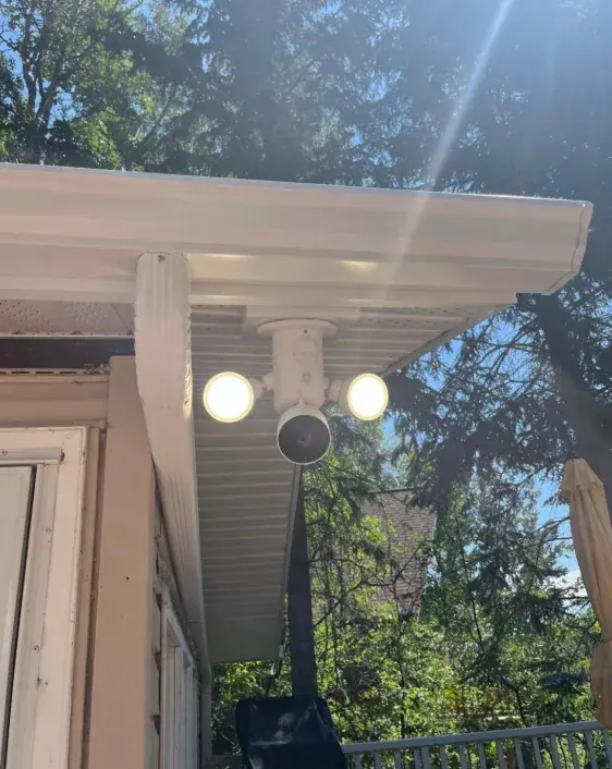 Twin-head LED security floodlight with a camera mounted under a white porch soffit, both lamps lit, trees and sunlight behind