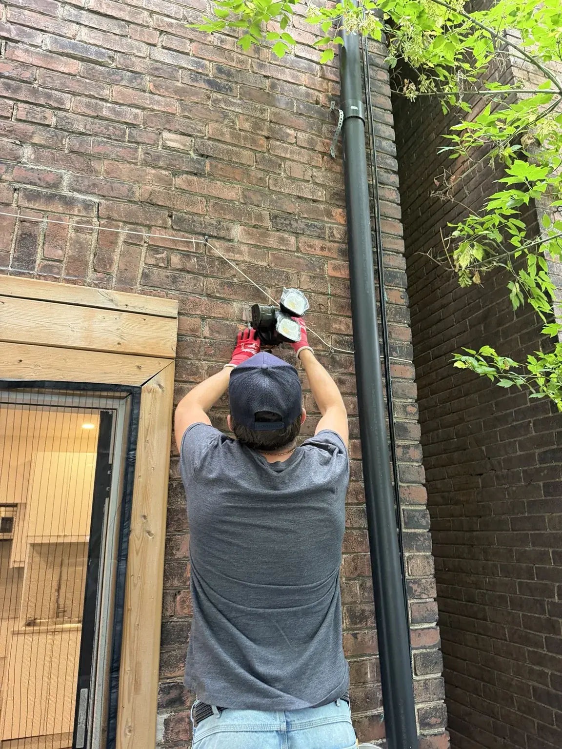 Electrician seen from behind, reaching up to mount a black wall light on a brick exterior wall beside a door with wood trim