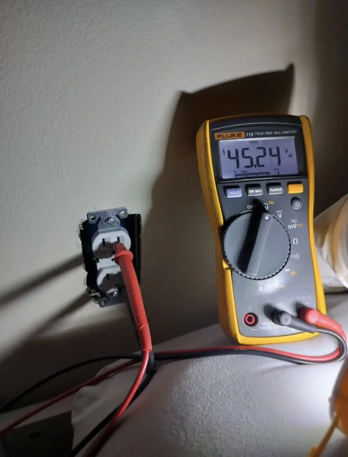 Digital multimeter standing on a counter with its probes in a receptacle whose cover plate has been removed; the display reads 45.24 volts AC