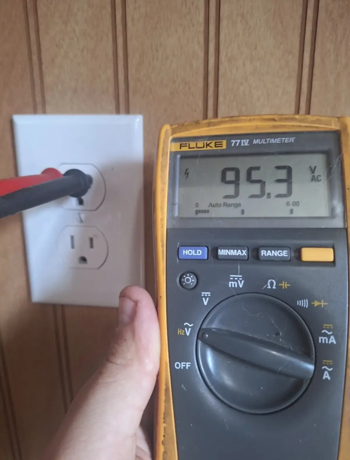Hand holding a digital multimeter in front of a wall receptacle in a wood-panelled room, one probe inserted; the display reads 95.3 volts AC