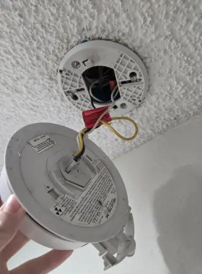 Smoke alarm unhooked from its ceiling mounting plate and hanging by its wiring harness below a textured ceiling with the box visible