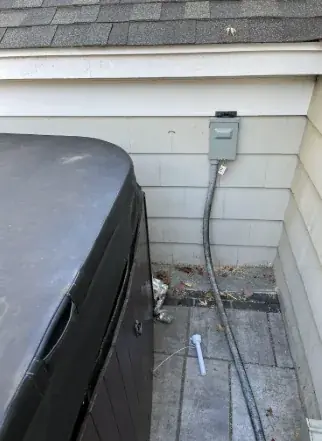 Grey electrical disconnect box mounted on the wall of a house beside a covered hot tub, with flexible conduit running down the wall and across the paving