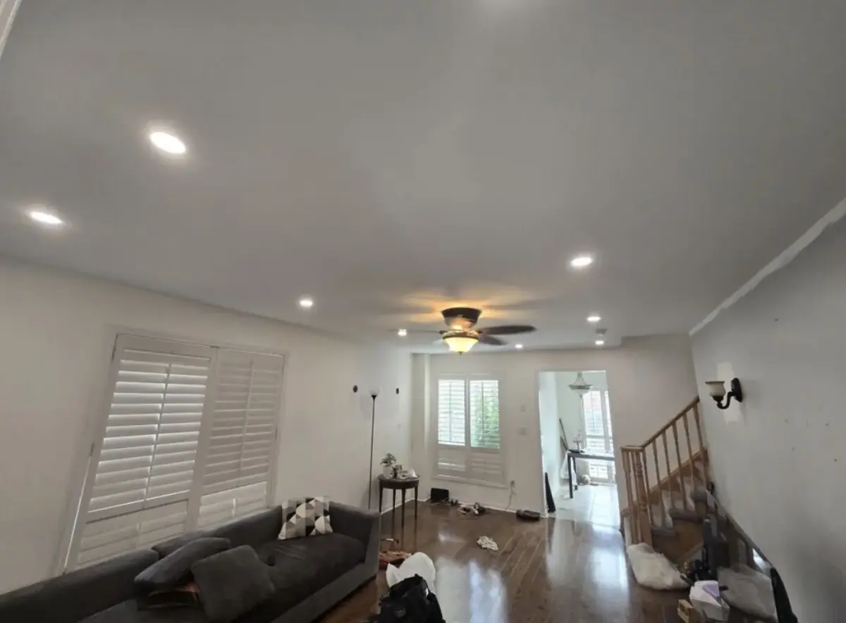 Living room with recessed pot lights across a white ceiling, a ceiling fan with a light, plantation shutters and a hardwood floor